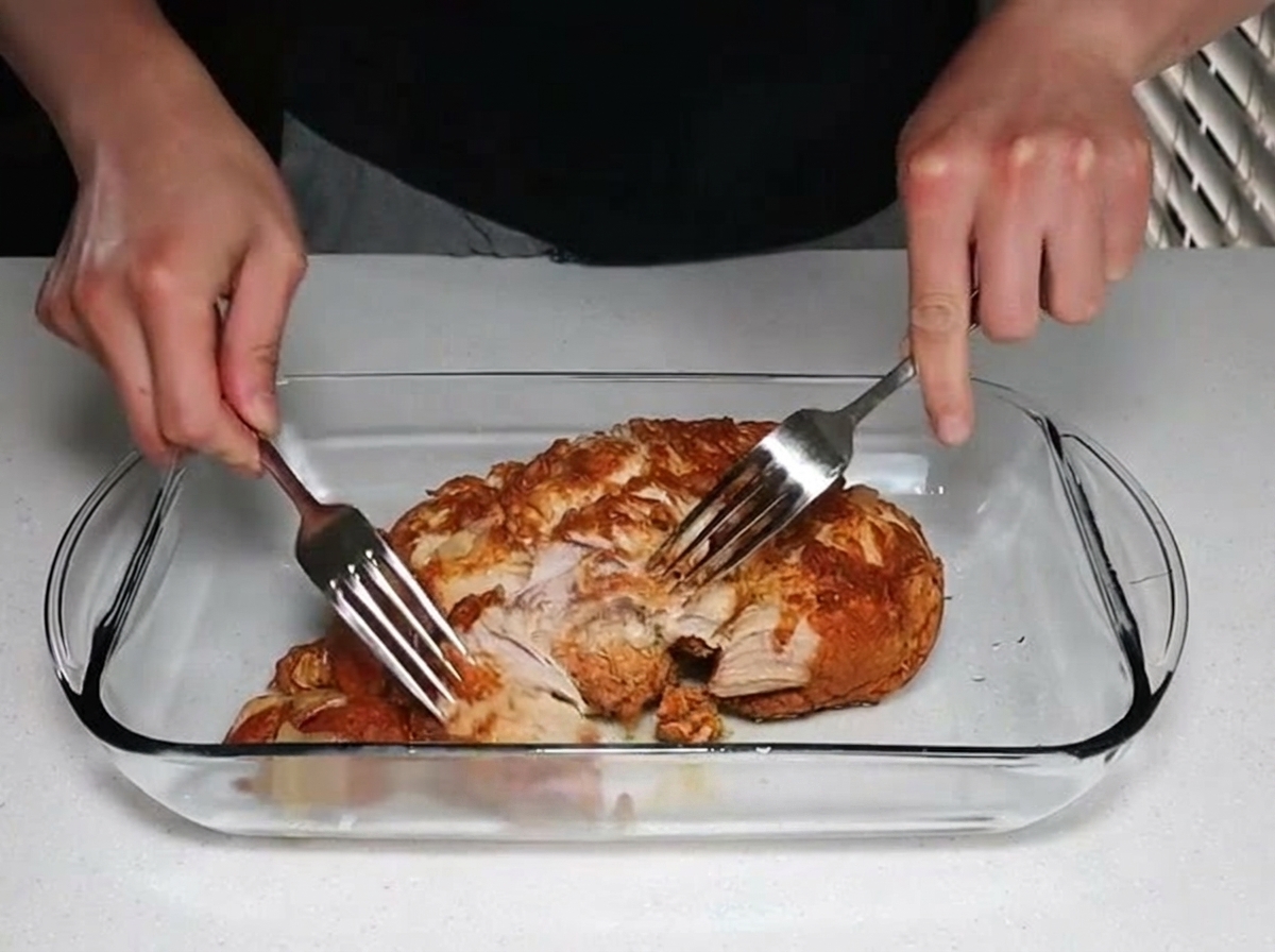Deux mains utilisant des fourchettes en acier inoxydable pour détacher et effilocher un morceau de porc cuit dans un plat en verre.