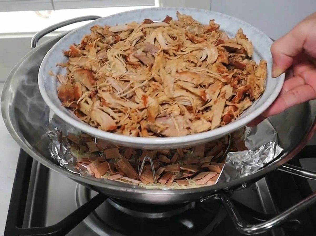 Une main déposant une assiette blanche remplie de porc effiloché sur une grille à lintérieur dun wok au-dessus de copeaux de bois fumants.