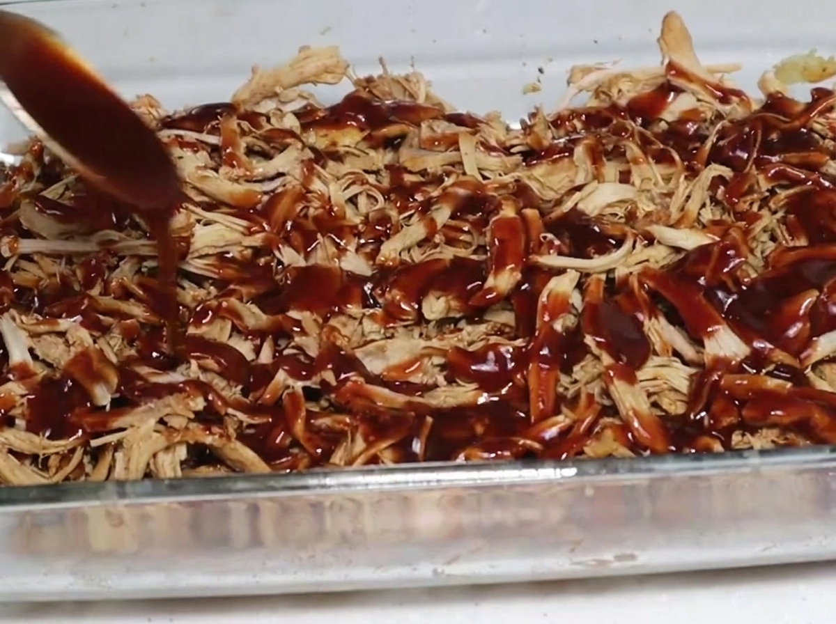 Une cuillérée de sauce barbecue épaisse et foncée nappant une couche de porc effiloché fumé dans un plat de cuisson.