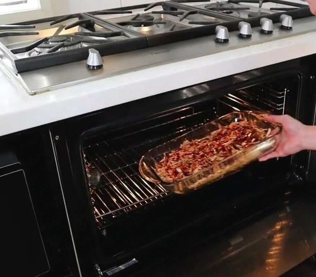Une personne glissant un plat en verre rempli de porc effiloché en sauce sur la grille centrale dun four chaud.