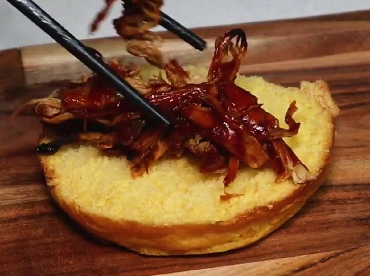 Une paire de baguettes déposant du porc effiloché juteux enrobé de sauce barbecue foncée sur la surface dorée dun demi-pain à burger grillé.
