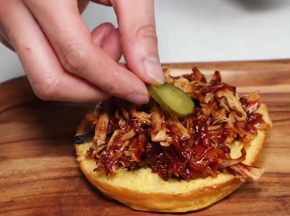 Une main déposant une rondelle de cornichon sur un dôme de porc effiloché barbecue dans un pain à burger grillé.