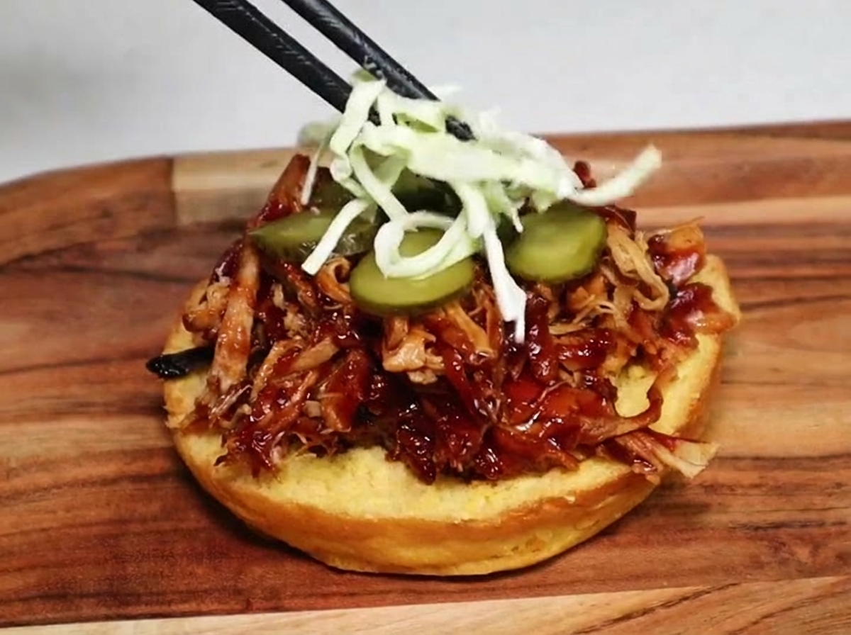 Des baguettes déposant de la salade de chou blanc émincé sur des rondelles de cornichons et du porc effiloché dans un pain à burger.