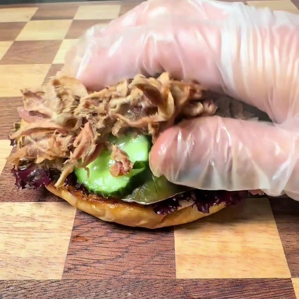 Une main portant un gant en plastique transparent déposant du porc effiloché tendre sur une base de burger garnie de laitue et de rondelles de cornichon.