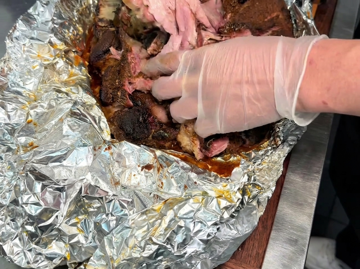 Des mains gantées détachant et effilochant un gros morceau de porc fumé tendre et cuit sur une surface recouverte daluminium.