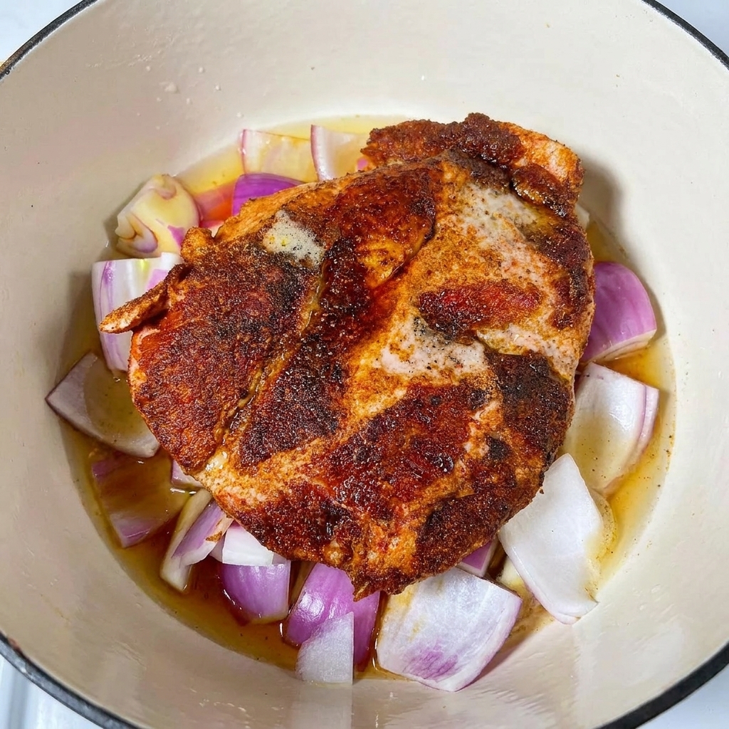 Liquides de braisage et bière versés dans une cocotte autour dune épaule de porc assaisonnée sur un lit doignons rouges.