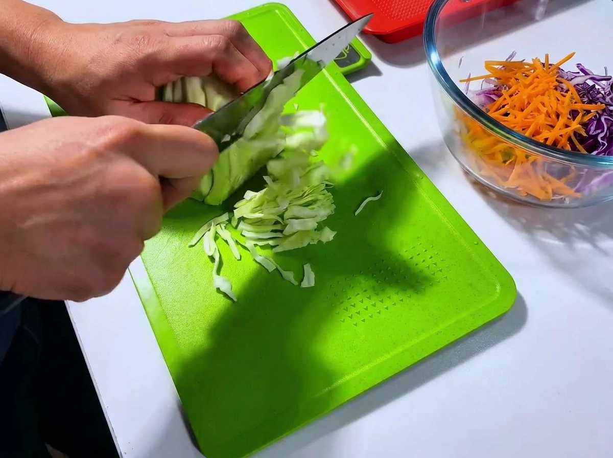 Un chef coupant une tête de chou vert en fines lanières avec un grand couteau sur une planche à découper vert vif.