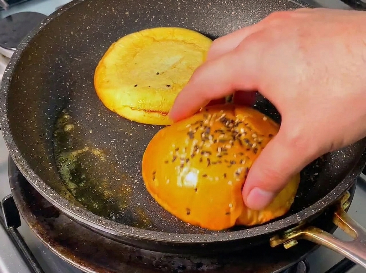 Une main retournant une moitié de pain à burger dorée dans une poêle chaude avec du beurre fondu.