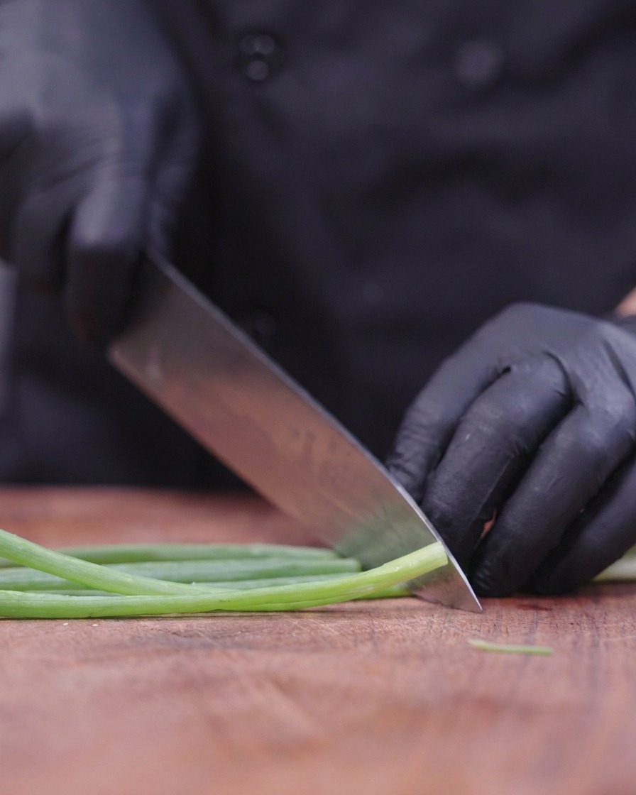 Un chef portant des gants noirs utilise un grand couteau pour émincer finement des tiges doignons verts sur une planche en bois.