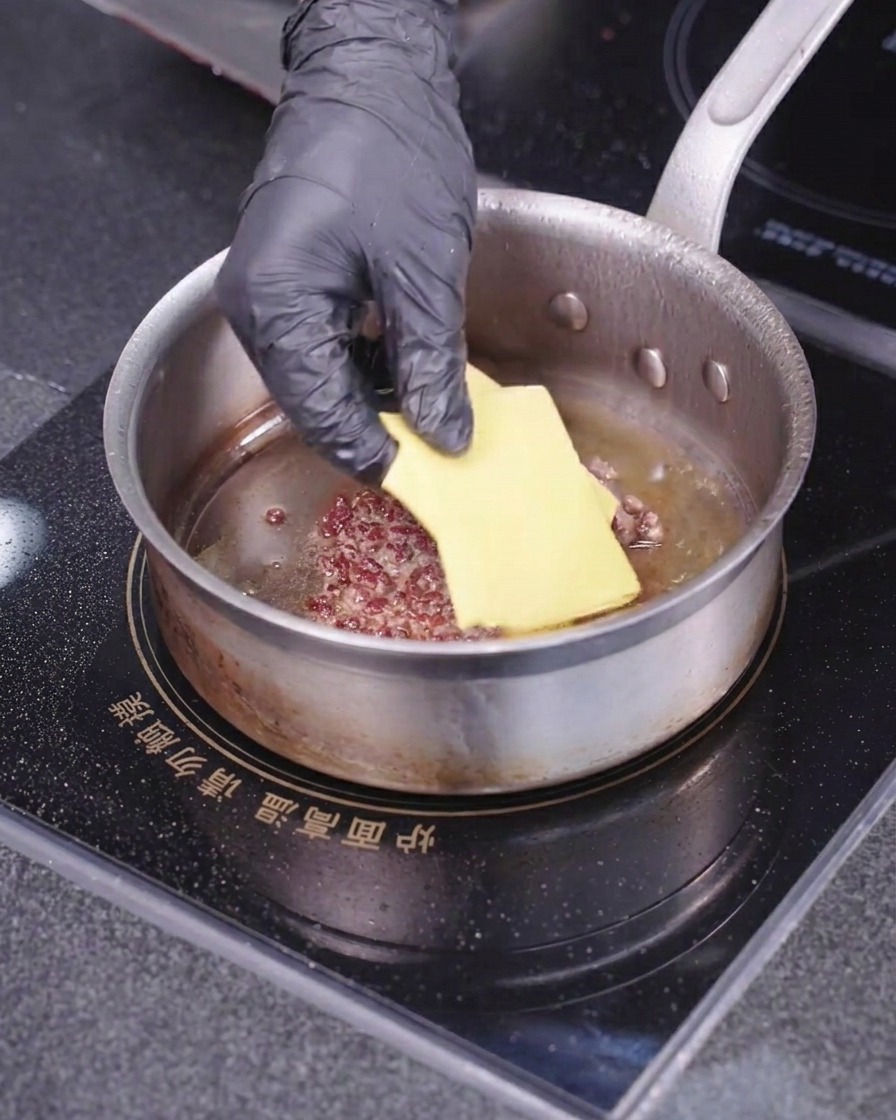 Une main gantée déposant une tranche carrée de fromage jaune sur un steak de viande en cuisson dans une poêle chaude.