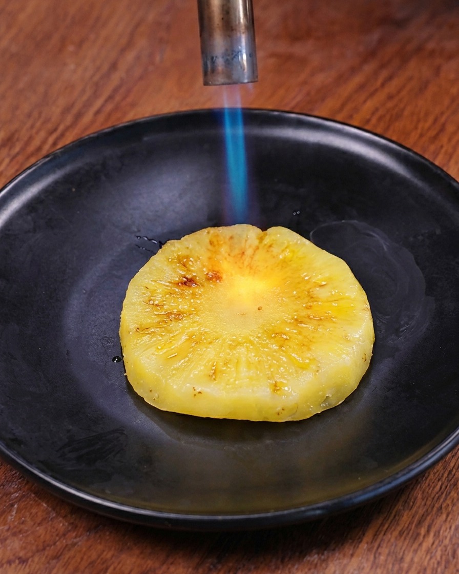 La flamme bleue dun chalumeau de cuisine venant dorer la surface dune tranche dananas frais sur une assiette sombre.