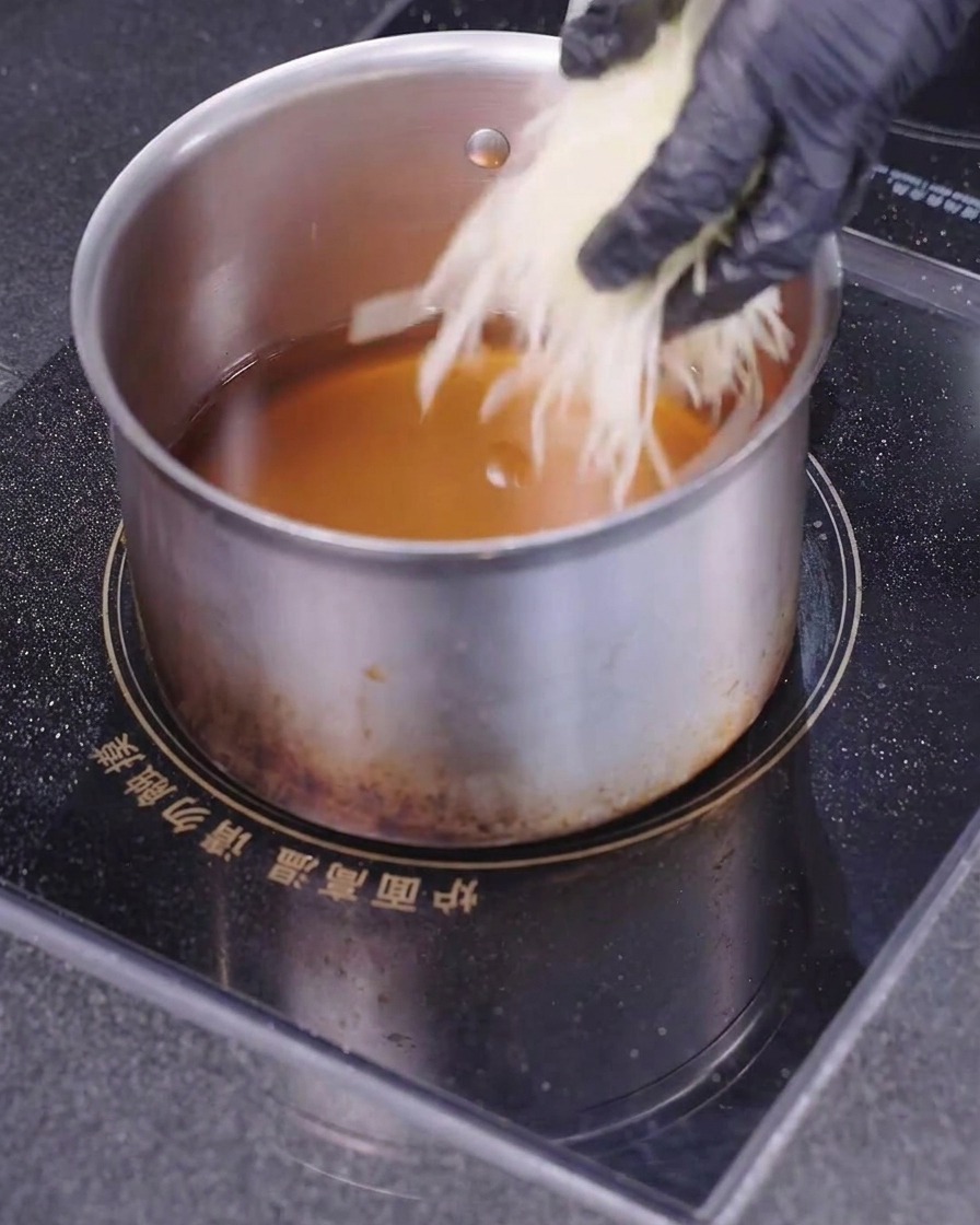 Une main portant un gant noir plongeant de fines lamelles de pommes de terre blanches dans une casserole remplie dhuile de friture chaude.