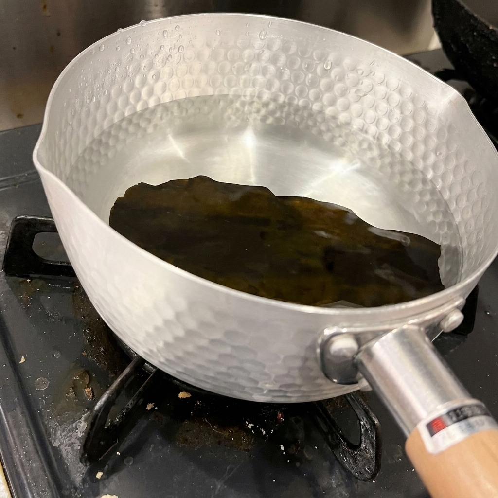 Un morceau dalgue kombu séchée et sombre repose au fond dune casserole en aluminium martelé remplie deau claire et froide.