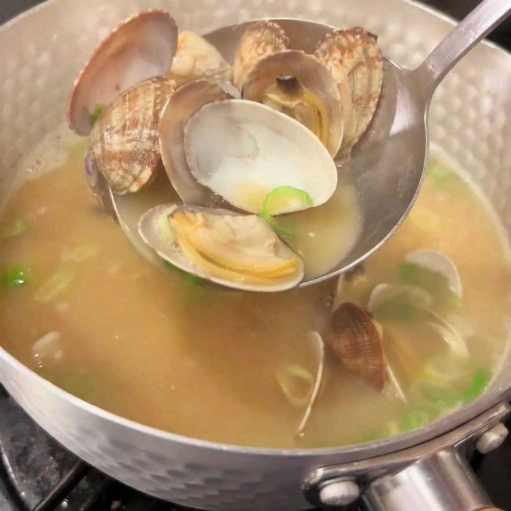Une louche en métal puise des palourdes ouvertes dans une casserole de soupe miso japonaise fumante, avec de vibrants oignons verts émincés mélangés au bouillon.