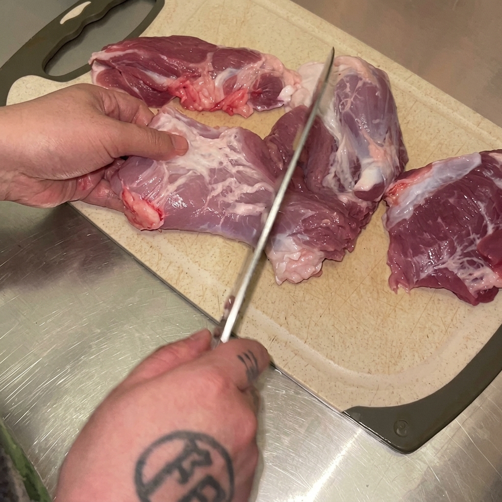 Mains utilisant un couteau pour couper de la cuisse de porc crue en morceaux sur une planche à découper.