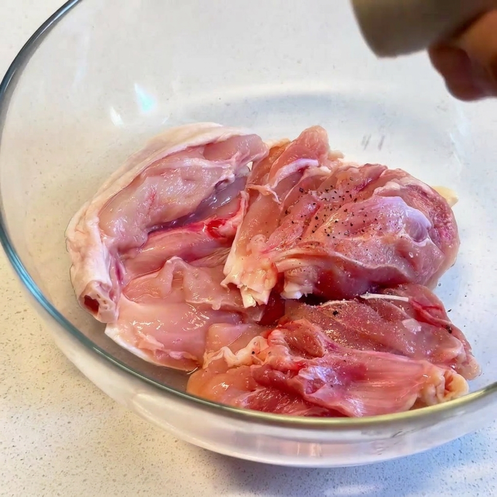 Cuisses de poulet crues dans un bol en verre transparent, assaisonnées de poivre noir fraîchement moulu avec un moulin.