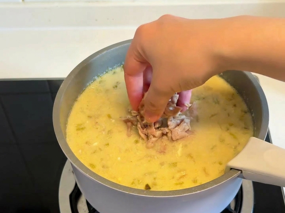 Une main plongeant des morceaux de poulet effiloché dans une marmite de soupe épaisse au citron.