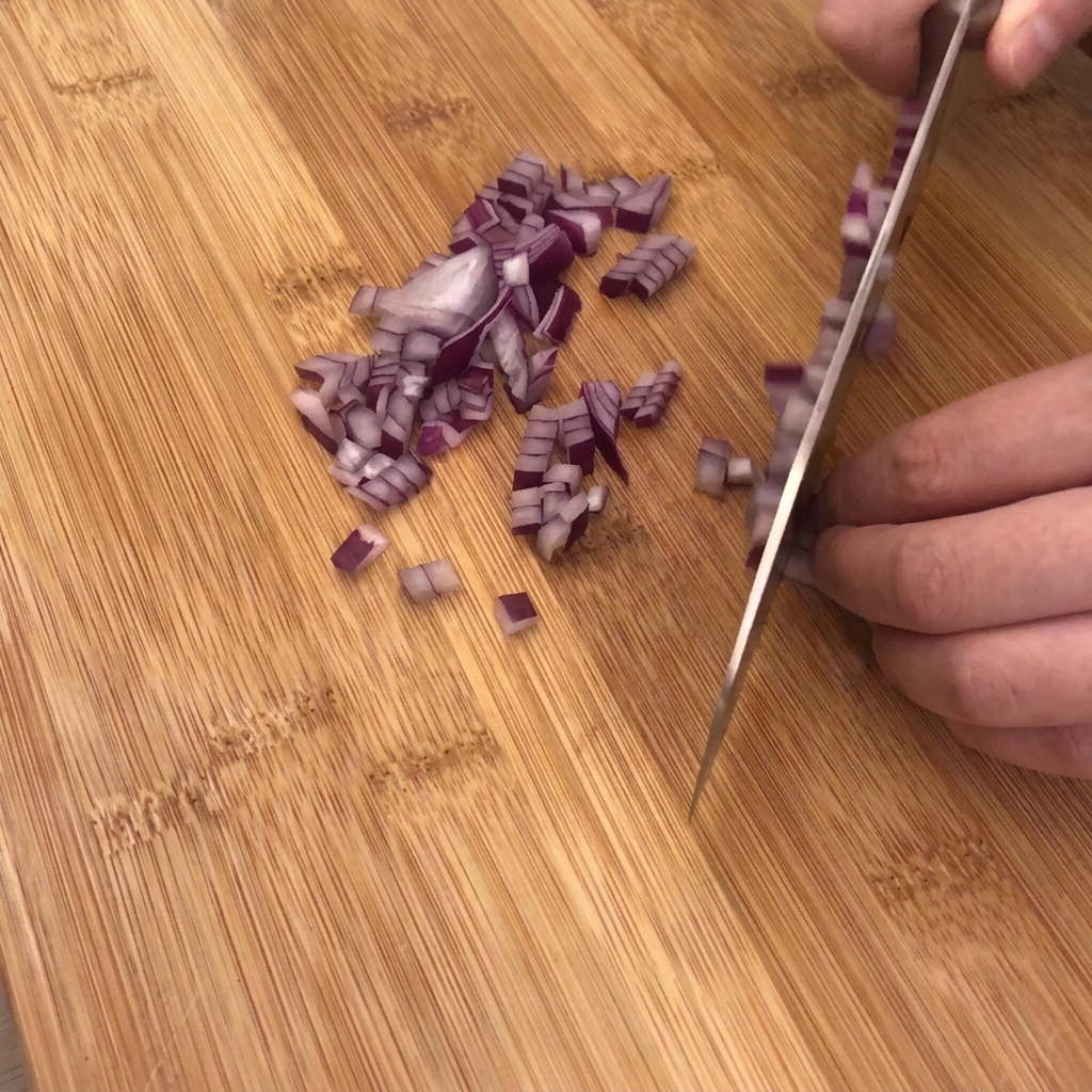 Un couteau de cuisine coupant en dés un oignon rouge sur une planche en bois, avec de lail et du bacon à proximité.