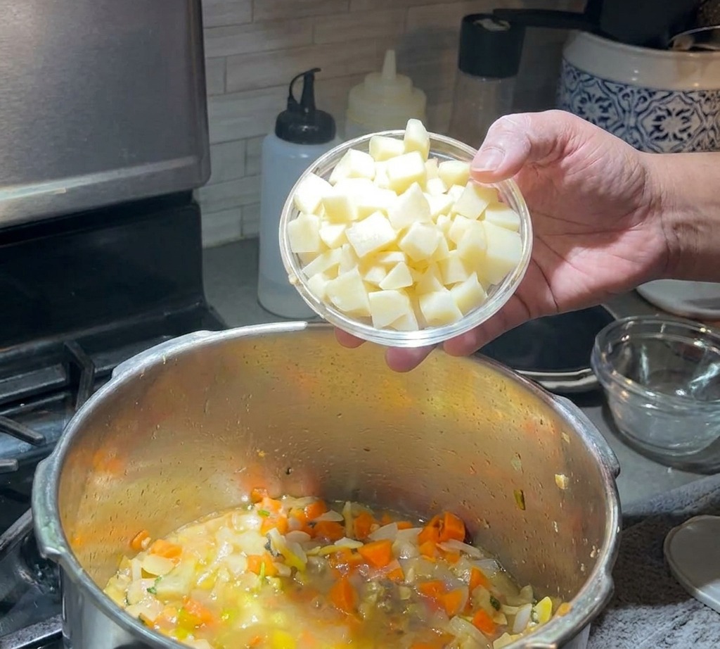 Une main versant un petit bol de pommes de terre en cubes dans une casserole de soupe mijotante remplie de carottes et doignons.