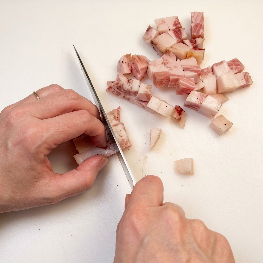 Main avec un couteau de chef coupant du bacon en dés sur une planche à découper.