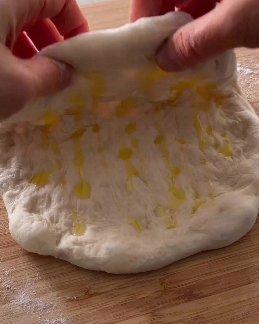 Mains pliant un morceau de pâte à pizza huilée en deux sur une surface en bois.