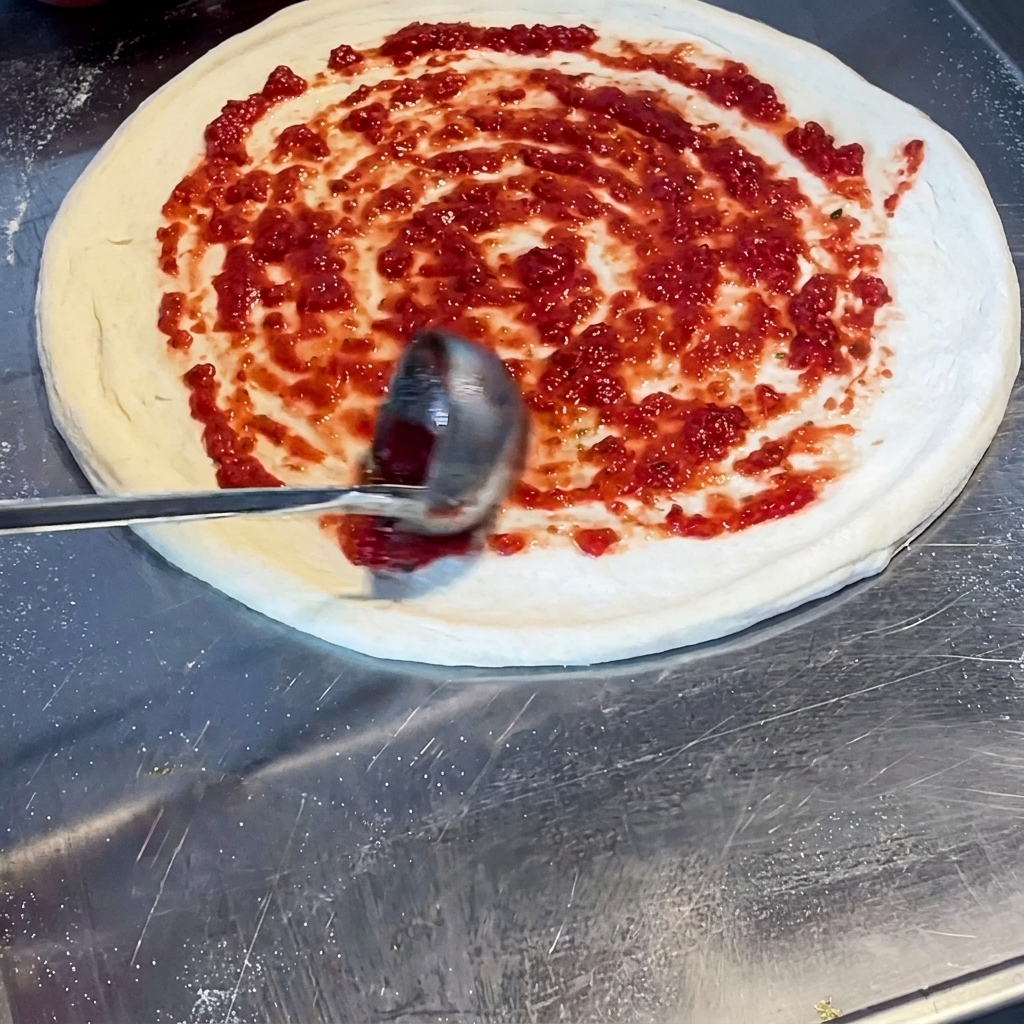 Une louche étalant une sauce tomate rouge vive en spirale sur la pâte à pizza crue.