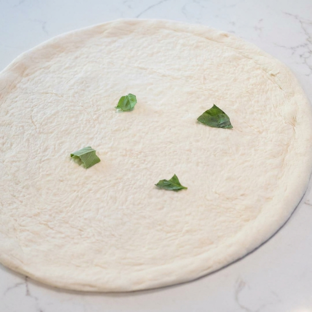 Une base de pâte à pizza crue étalée, garnie de feuilles de basilic frais dispersées.