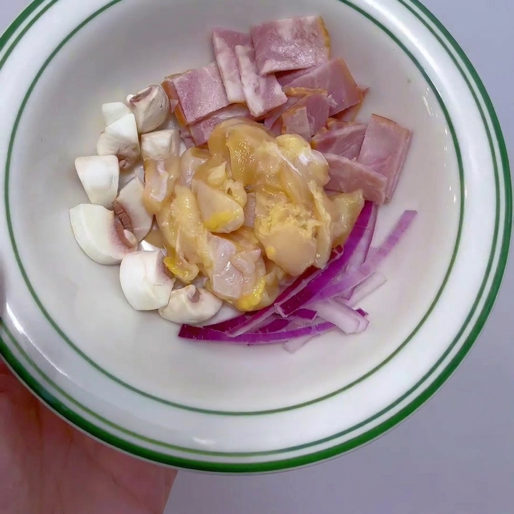 Un petit bol blanc contenant du blanc de poulet cru en dés, des tranches de bacon, des champignons de Paris en quartiers et de loignon rouge émincé.