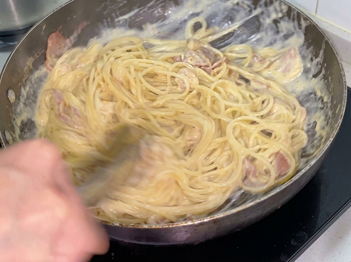Des spaghettis crémeux mélangés au bacon et au poulet à laide de pinces en métal dans une poêle.