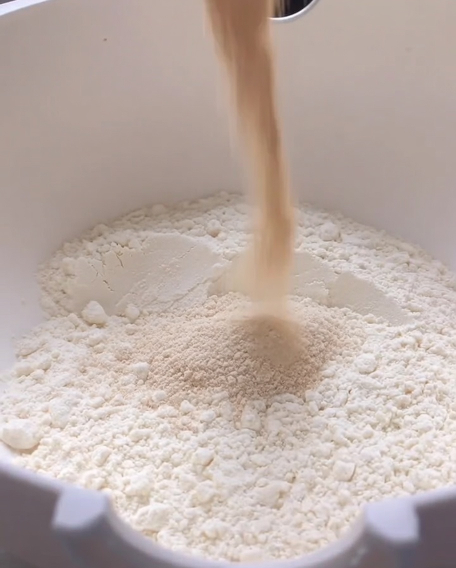Versement de levure sèche dans un grand bol blanc rempli de farine.