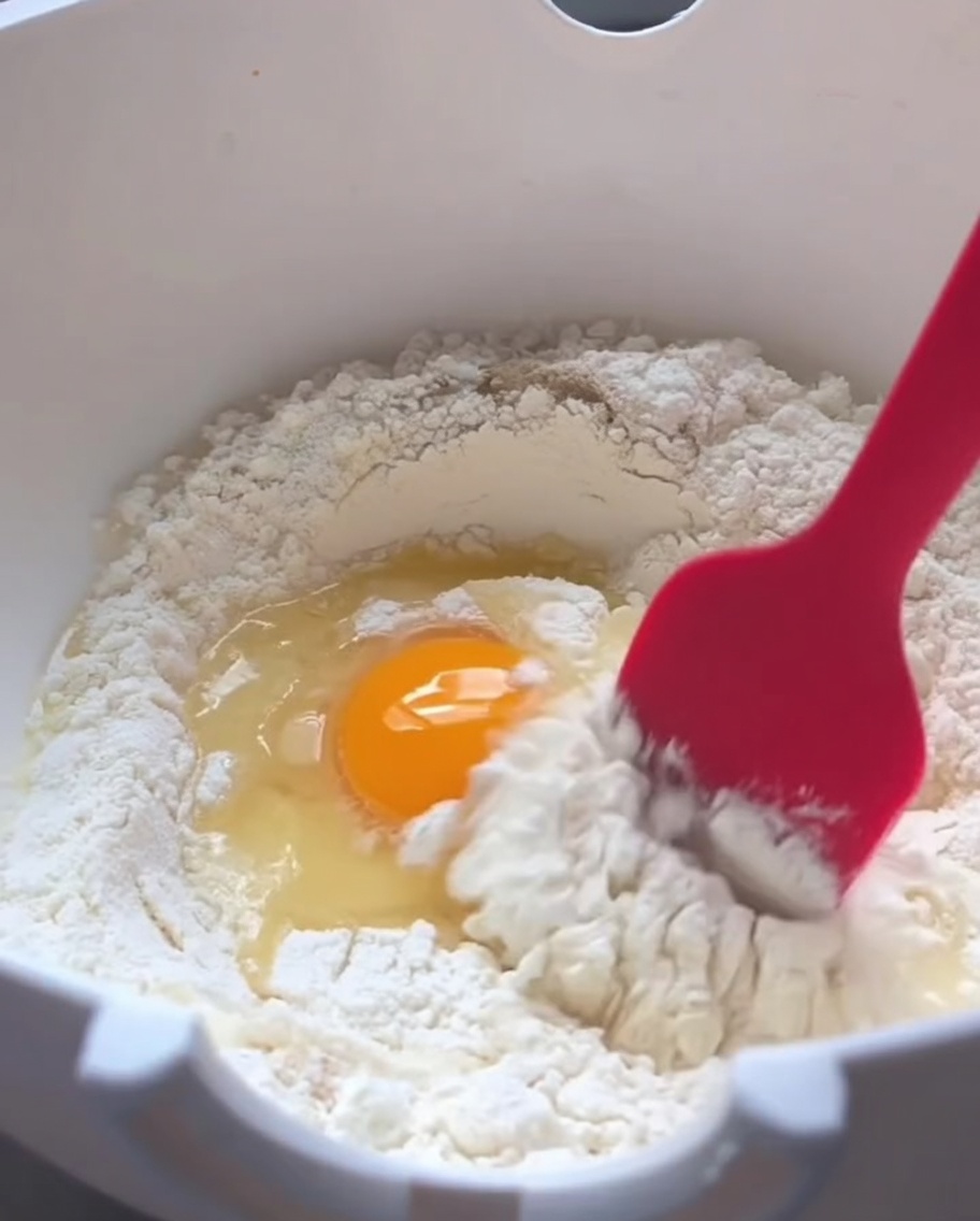 Une spatule en silicone rouge mélangeant de la farine, un œuf cru et de leau tiède dans un bol blanc.
