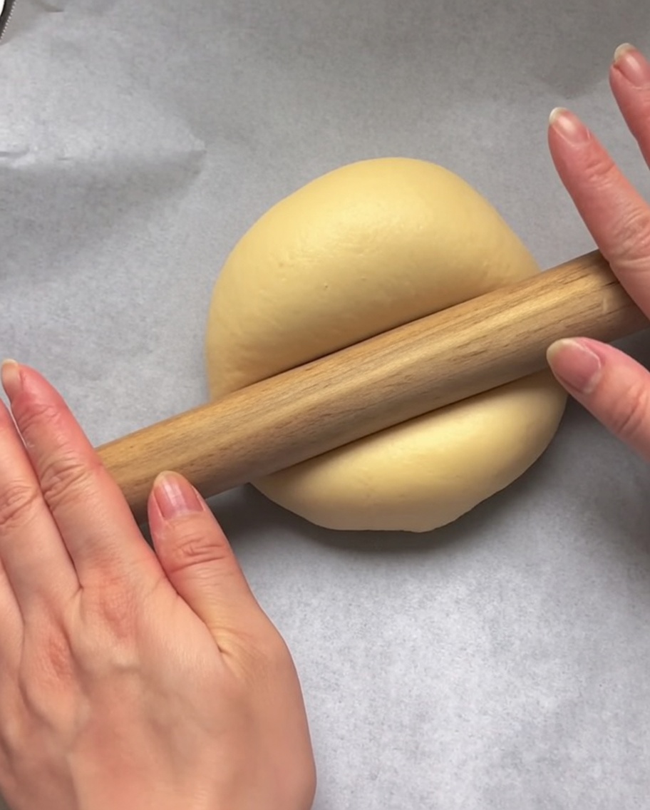 Des mains utilisant un rouleau à pâtisserie en bois pour aplatir une boule de pâte crue en forme de cercle sur du papier cuisson blanc.