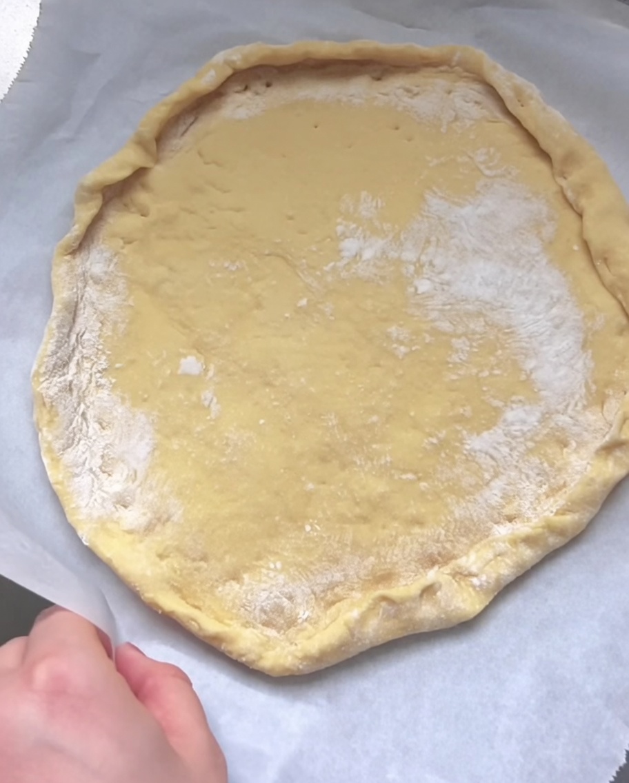 Une main repliant et pinçant le bord de la pâte aplatie pour former une croûte surélevée sur le papier cuisson.