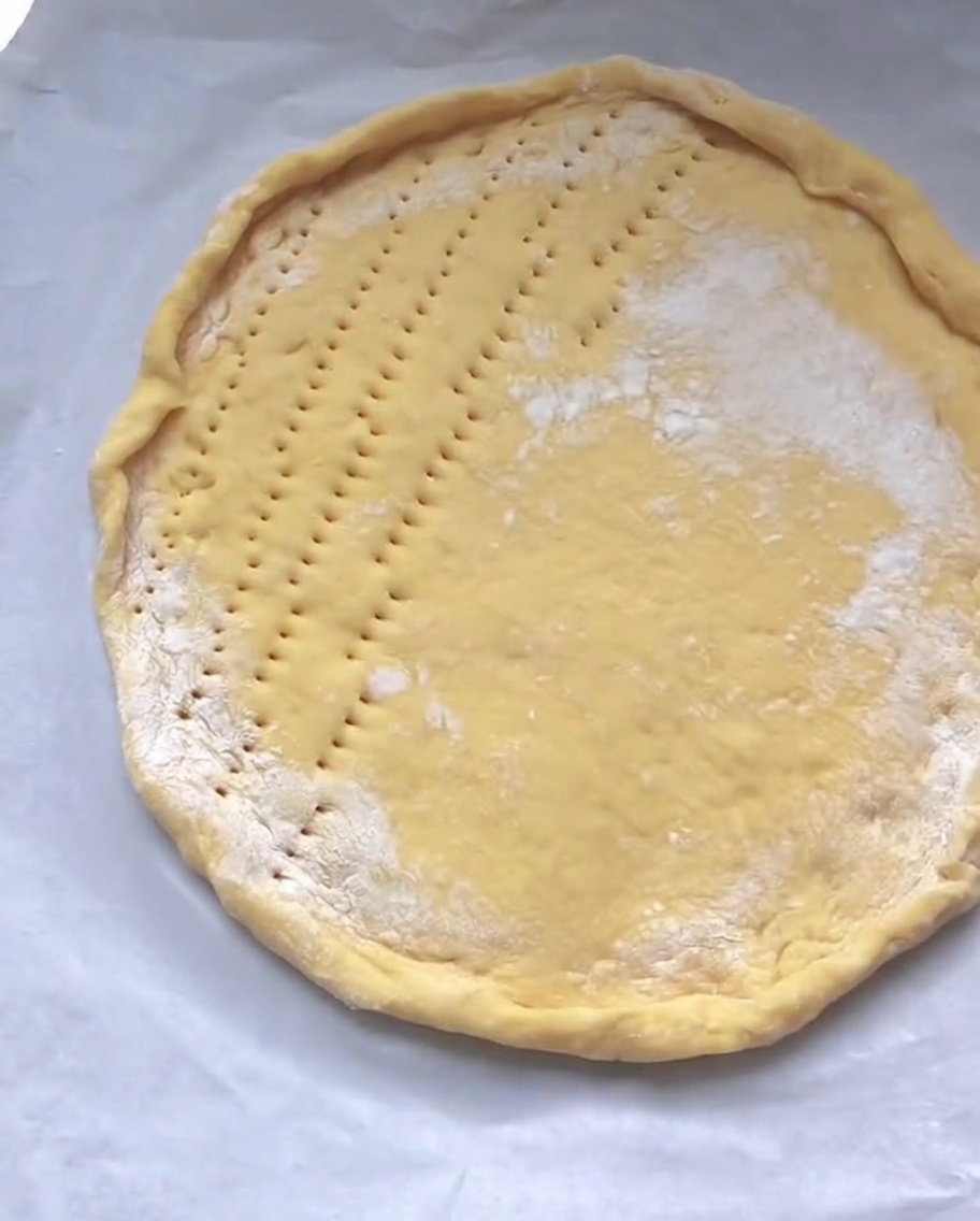 Une pâte à pizza circulaire crue sur du papier cuisson avec de petits trous piqués uniformément sur sa surface centrale.