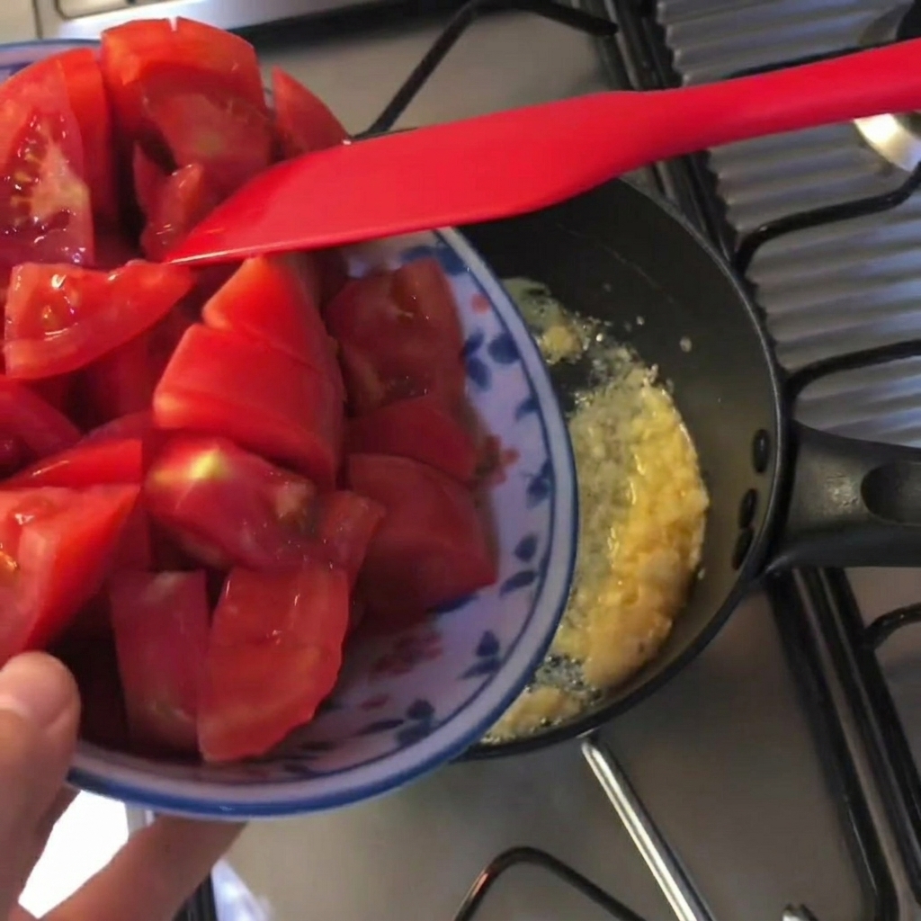 Des morceaux de tomates rouges fraîches ajoutés dans une poêle noire avec du beurre fondu et de lail haché.