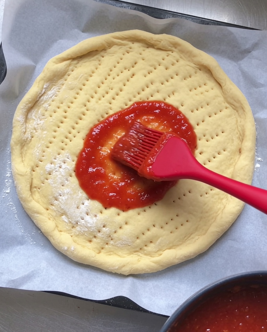 De la sauce tomate rouge étalée uniformément sur la base de la pâte à pizza crue et piquée.
