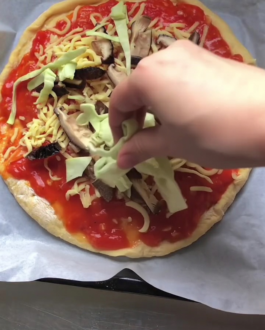 Une main déposant du chou vert cru émincé sur une pizza nappée de sauce tomate, de fromage et de champignons émincés.