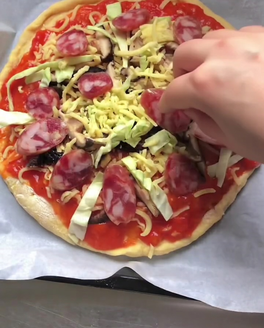 Une main plaçant des rondelles de saucisse italienne rouge sur une pizza crue garnie de sauce, de fromage et de légumes.