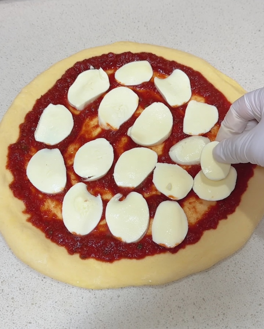 Main gantée déposant des tranches de mozzarella fraîche sur la base de la pizza.