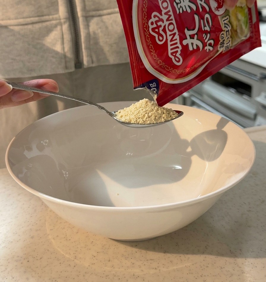 De la poudre de bouillon de poulet versée dun sachet dans une cuillère au-dessus dun grand bol de service blanc.