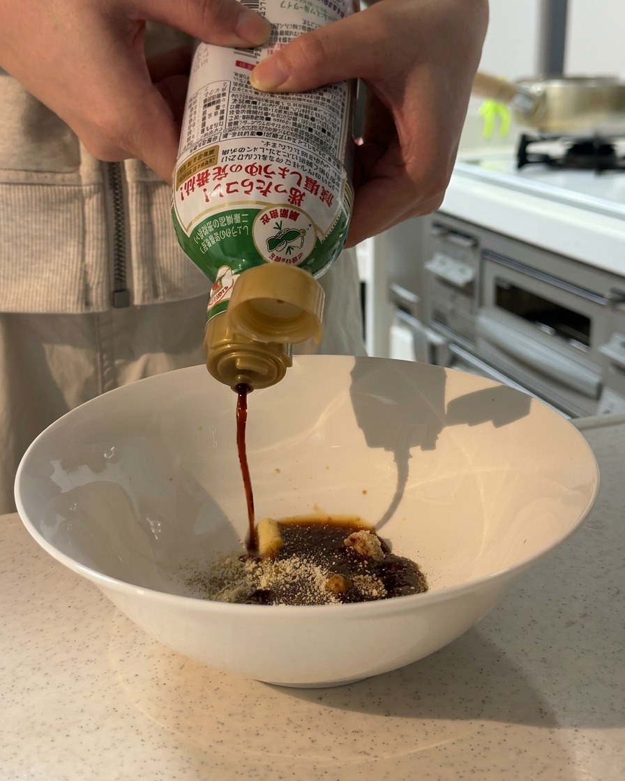Des mains versant de la sauce soja à teneur réduite en sel depuis une bouteille dans un bol blanc contenant des assaisonnements secs et de la pâte.