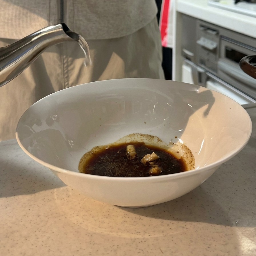 De leau bouillante versée depuis une bouilloire en acier inoxydable dans un bol blanc contenant un mélange de sauce soja et dassaisonnements sombres.