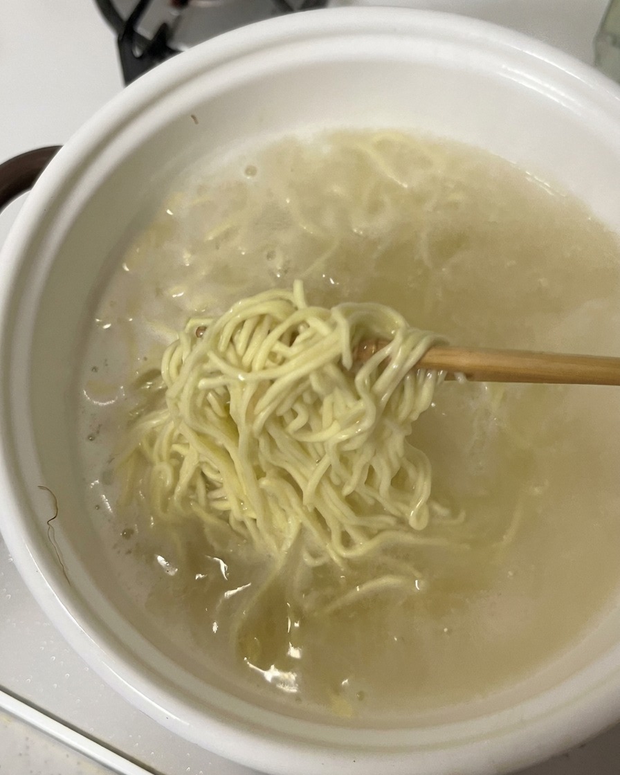 Des baguettes soulevant une portion de nouilles ramen cuites hors dune casserole blanche remplie deau bouillante.
