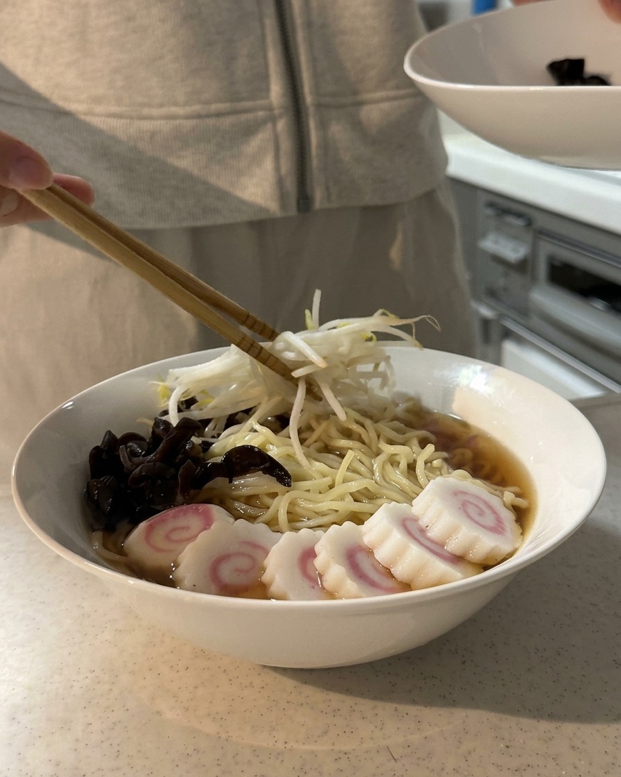 Des baguettes plaçant un tas de pousses de soja blanchies sur un bol de ramen déjà garni de champignons oreilles de bois et de gâteaux de poisson en spirale.