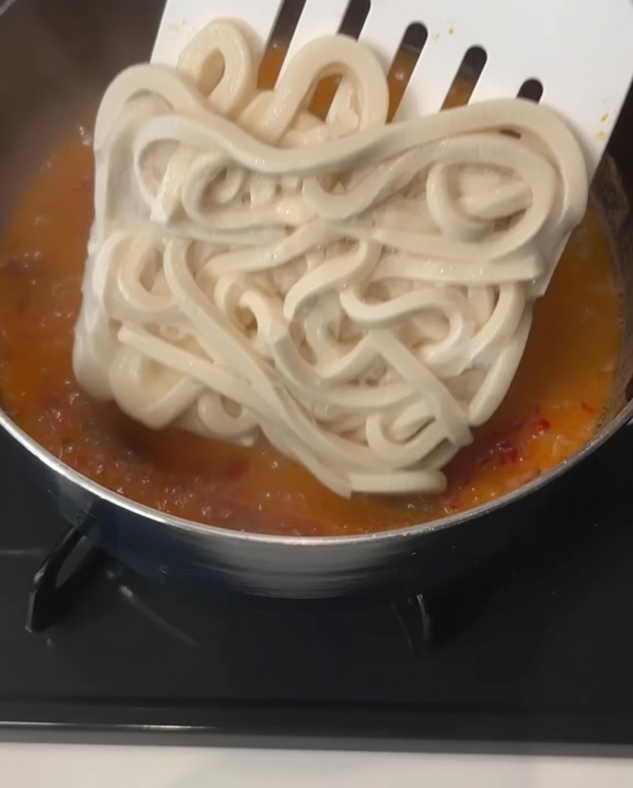 Un bloc carré de nouilles udon épaisses surgelées déposé par une spatule dans un bouillon crémeux orange frémissant.