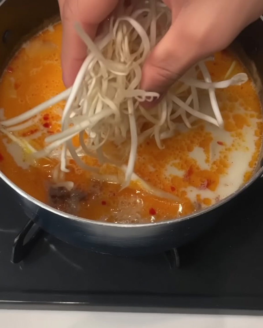 Une main jetant une poignée de pousses de soja blanches fraîches dans une casserole de soupe de nouilles crémeuse.