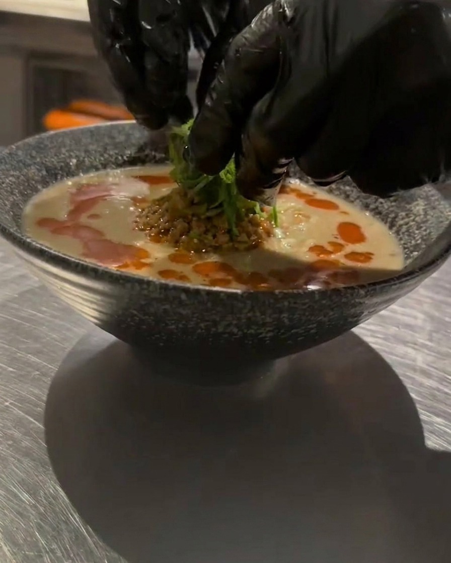 Mains gantées plaçant délicatement des oignons verts émincés sur un bol de Tan Tan Ramen chaud comme garniture finale.