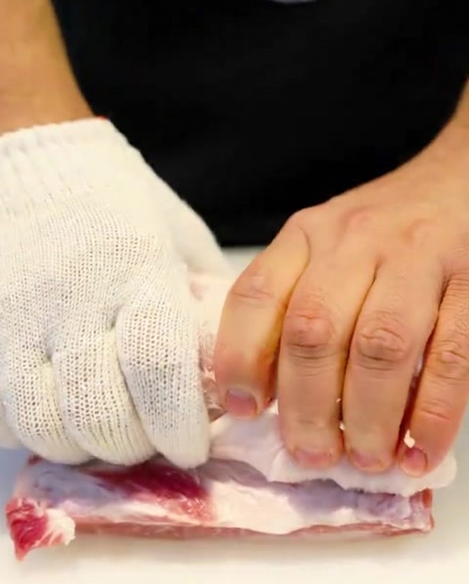 Un rouleau de poitrine de porc crue attaché serré avec de la ficelle de cuisine par des mains portant des gants de protection.
