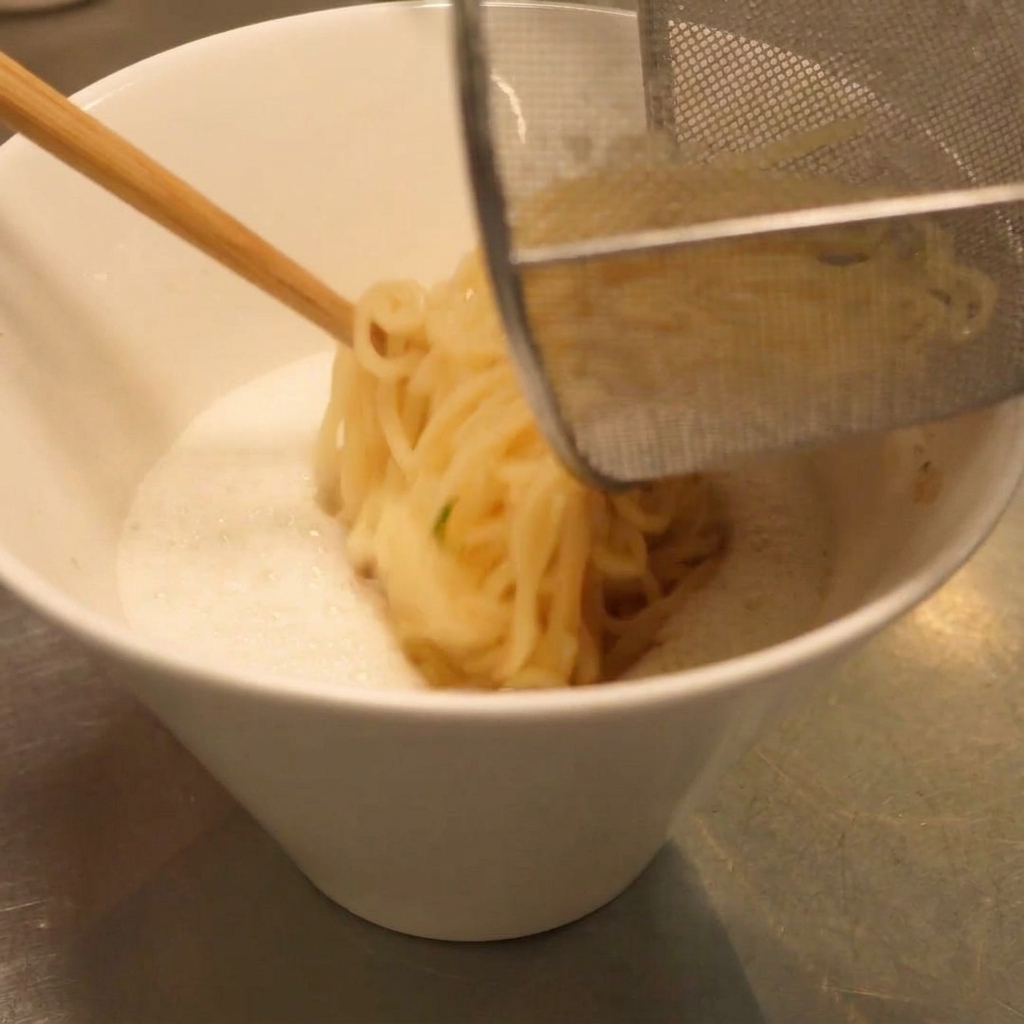 Des nouilles ramen égouttées transférées dans le bol de soupe crémeuse à laide dun panier de cuisine.