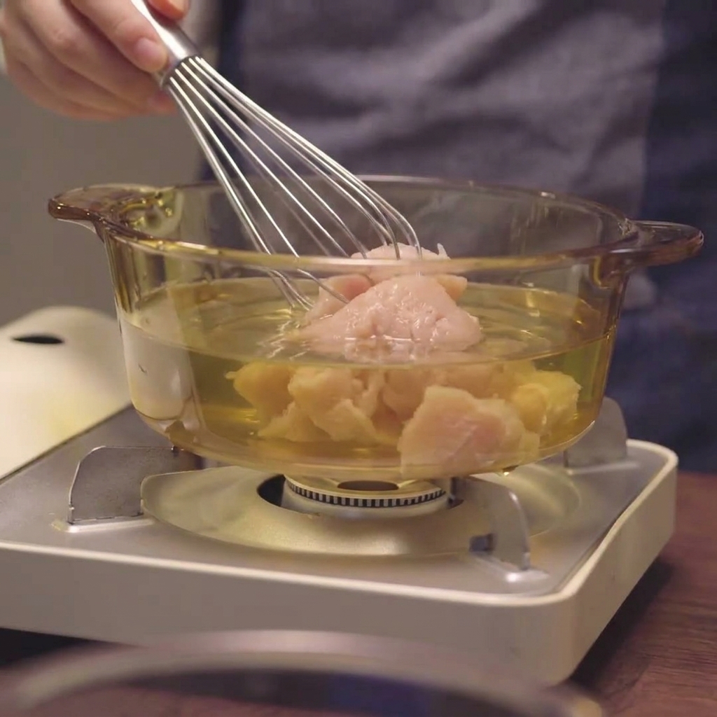 Un fouet mélangeant du blanc de poulet haché dans le liquide dune casserole transparente.